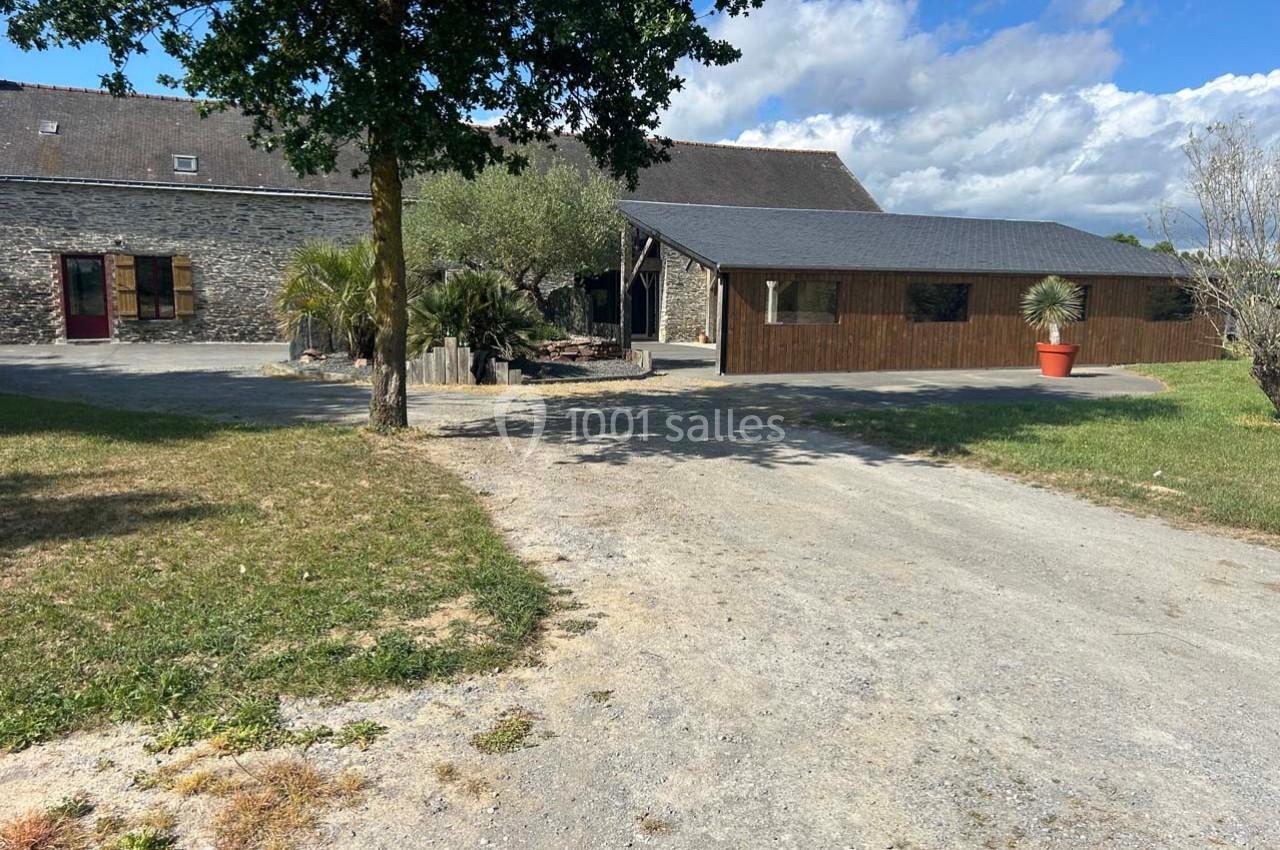 Bâtisse en pierre avec toit en ardoise, entourée de végétation et d'une cour en gravier sous un ciel partiellement nuageux.