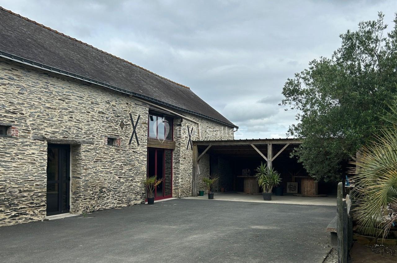 Bâtiment en pierre avec une grande baie vitrée, une terrasse couverte et des plantes, sous un ciel nuageux.
