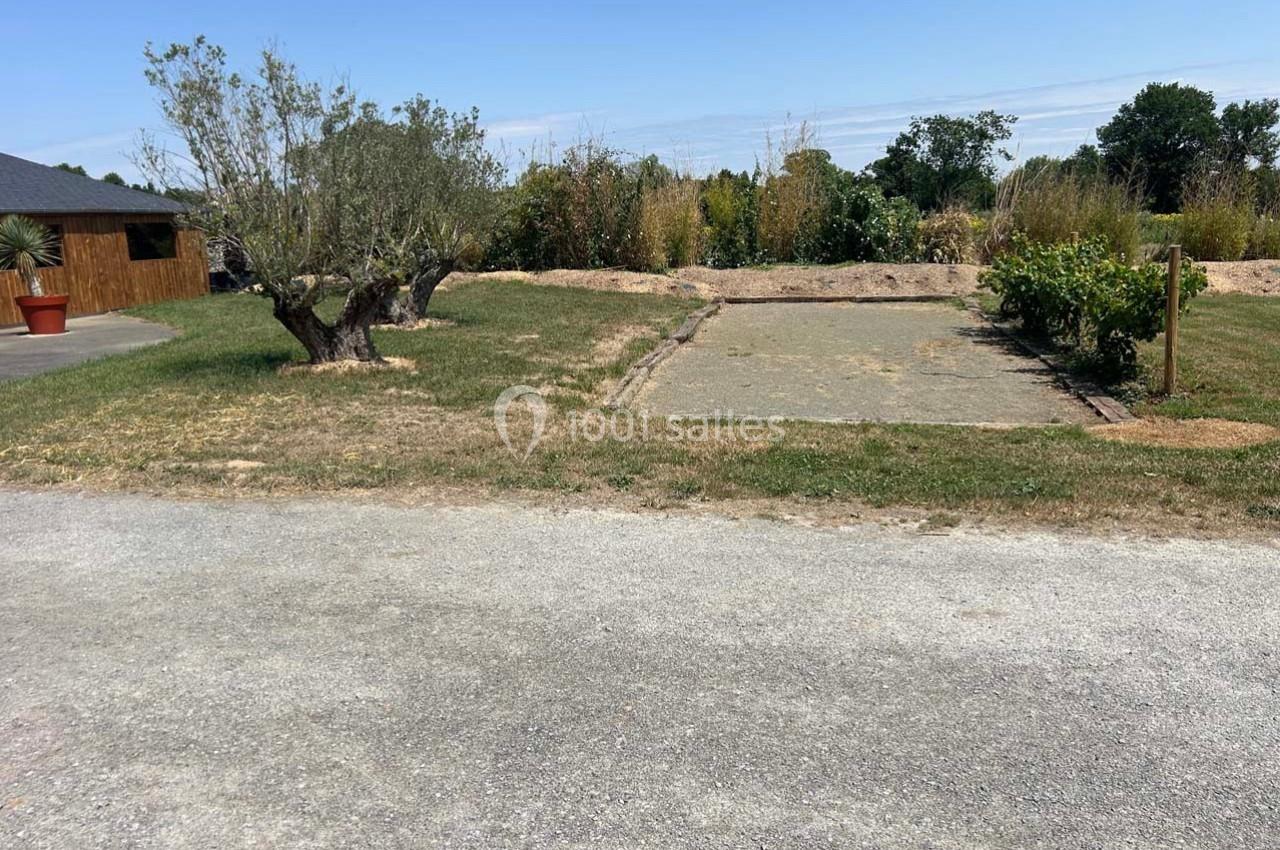 Terrain de pétanque en gravier entouré de végétation, avec un olivier et un bâtiment en arrière-plan.