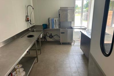 Cuisine professionnelle avec plans de travail en inox, évier, lave-vaisselle industriel et vue sur l'extérieur.