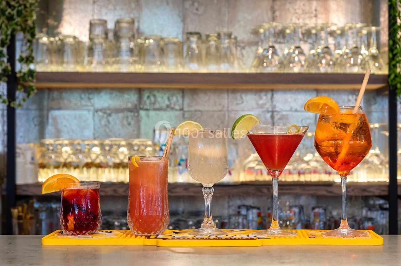 Cinq cocktails variés alignés sur un bar, avec des verres décorés de fruits et un fond de verres empilés.