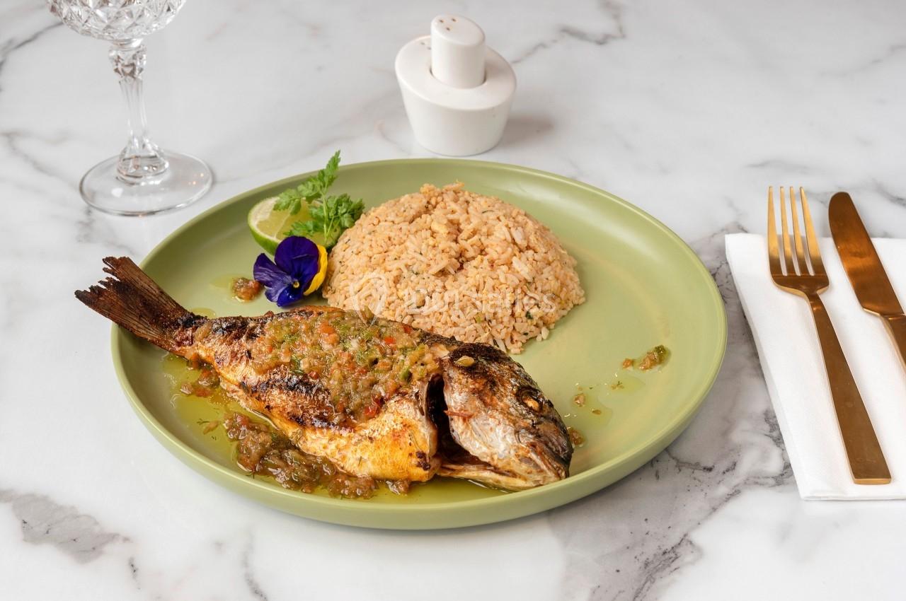 Poisson grillé avec sauce, servi avec du riz et une garniture de citron et fleur comestible sur une assiette verte.