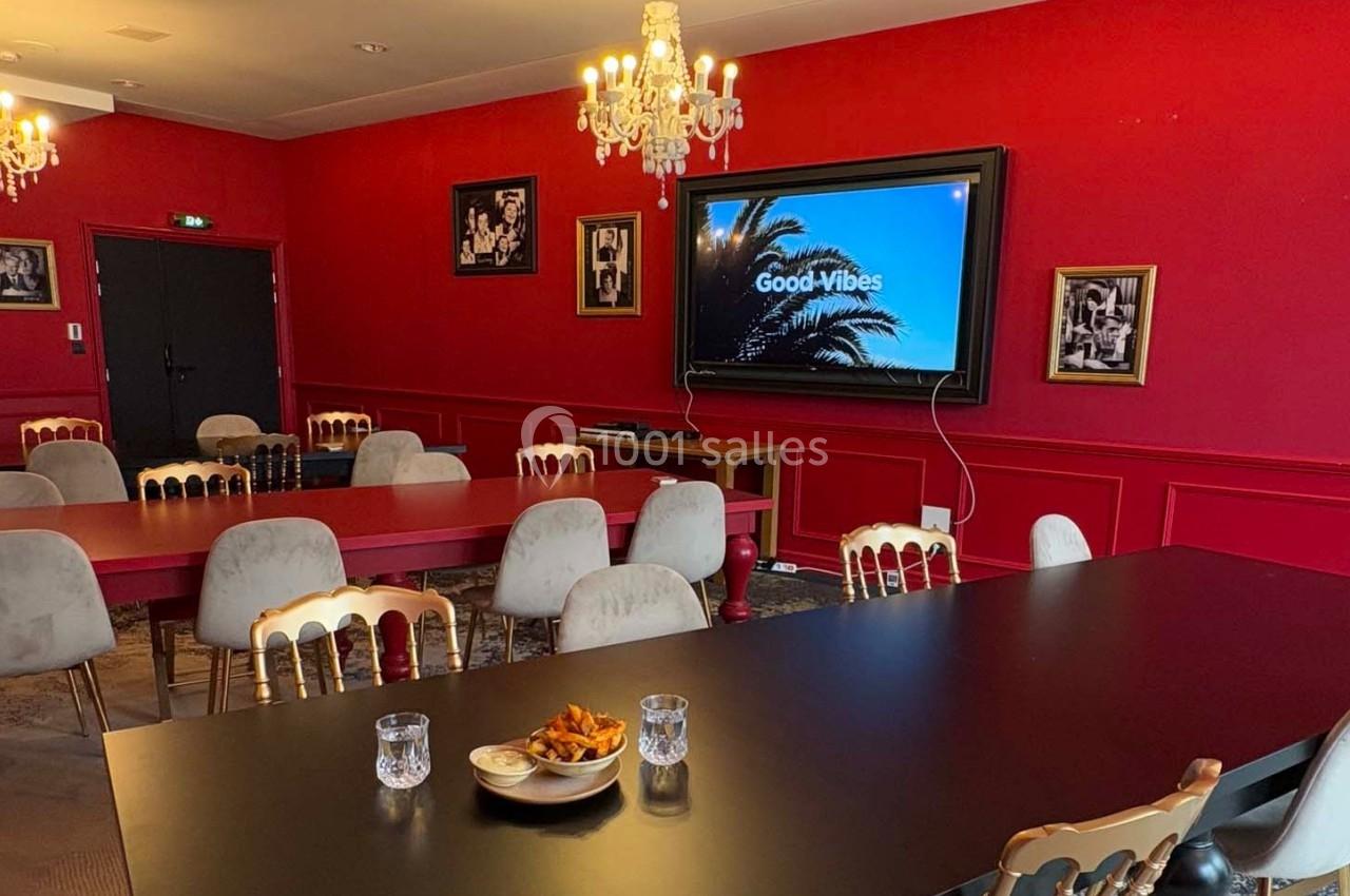 Salle de réunion avec murs rouges, tables noires, chaises dorées, écran affichant ’Good Vibes’ et lustres suspendus.