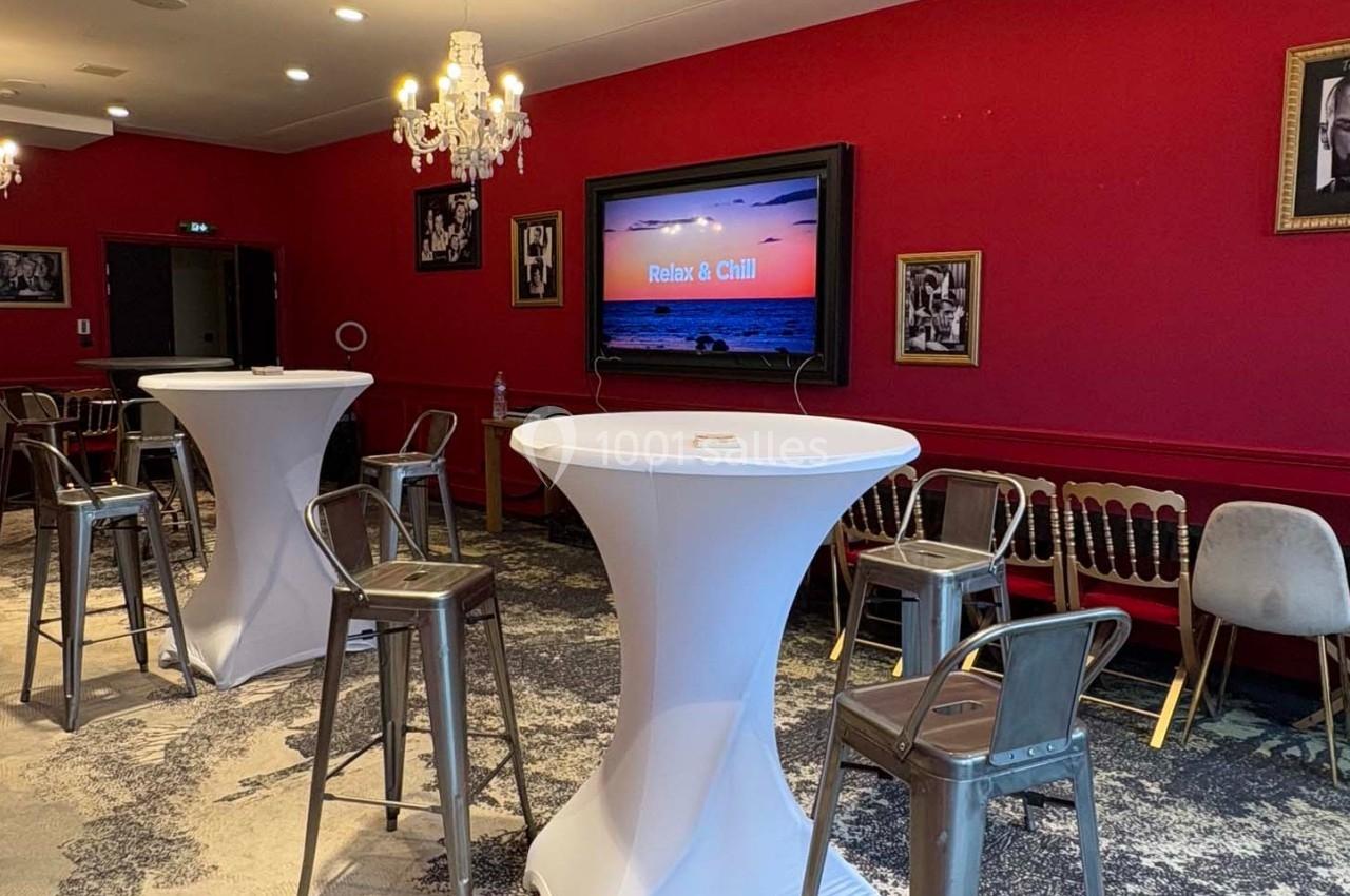 Salle avec murs rouges, tables hautes blanches, chaises métalliques et écran affichant ’Relax & Chill’.