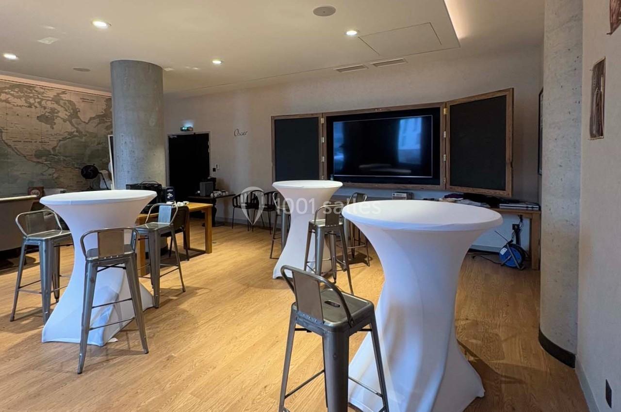 Salle lumineuse avec tables hautes drapées de blanc, chaises en métal, écran mural et décor sobre.