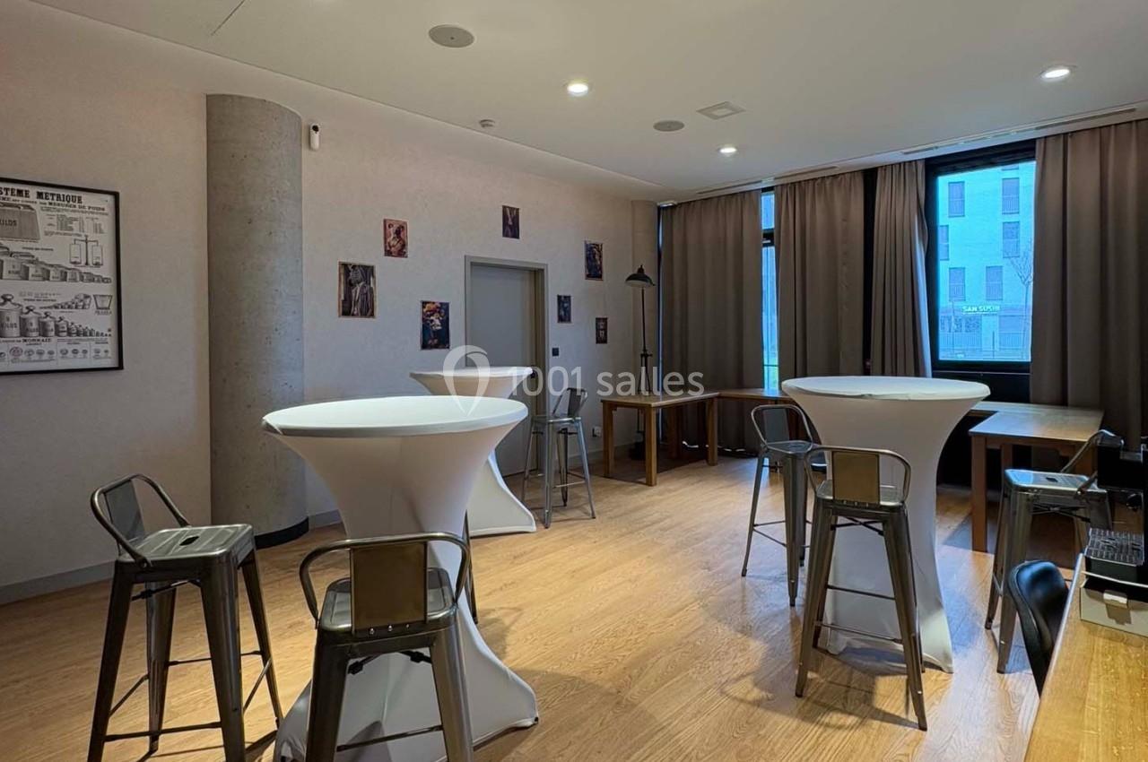 Salle lumineuse avec tables hautes, tabourets en métal, parquet en bois et grandes fenêtres avec rideaux.