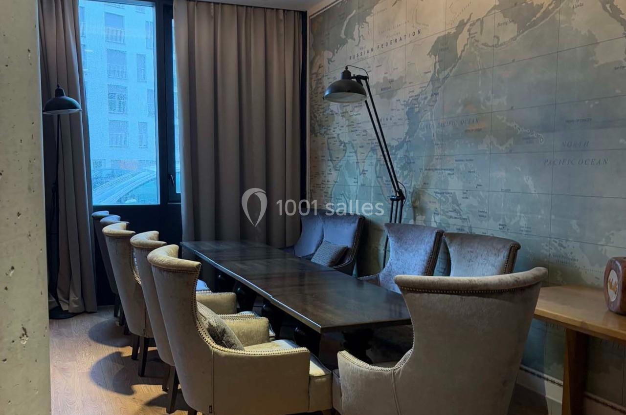 Salle de réunion avec une grande table en bois, entourée de fauteuils, éclairée par une fenêtre et une lampe murale.