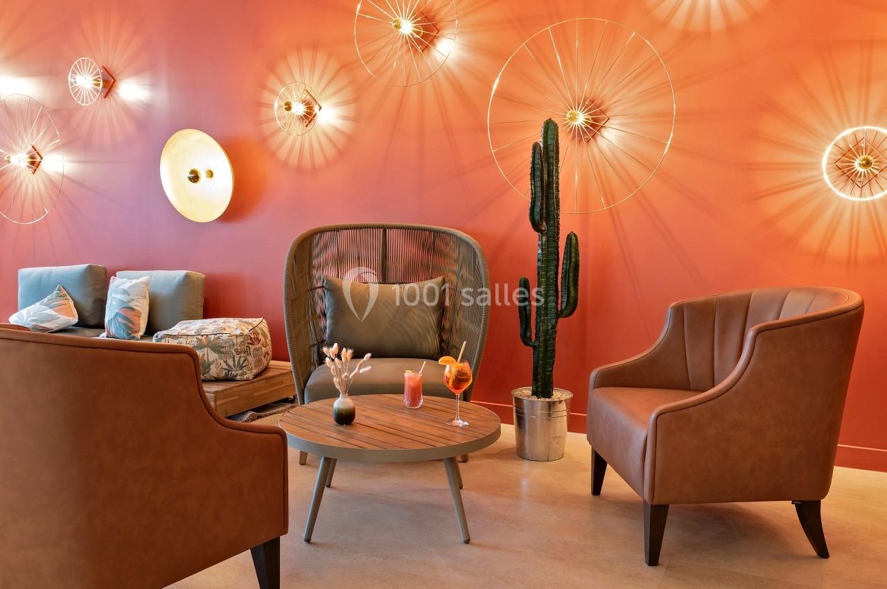 Espace salon avec fauteuils en cuir, table basse, cactus en pot et mur orange décoré de luminaires design.