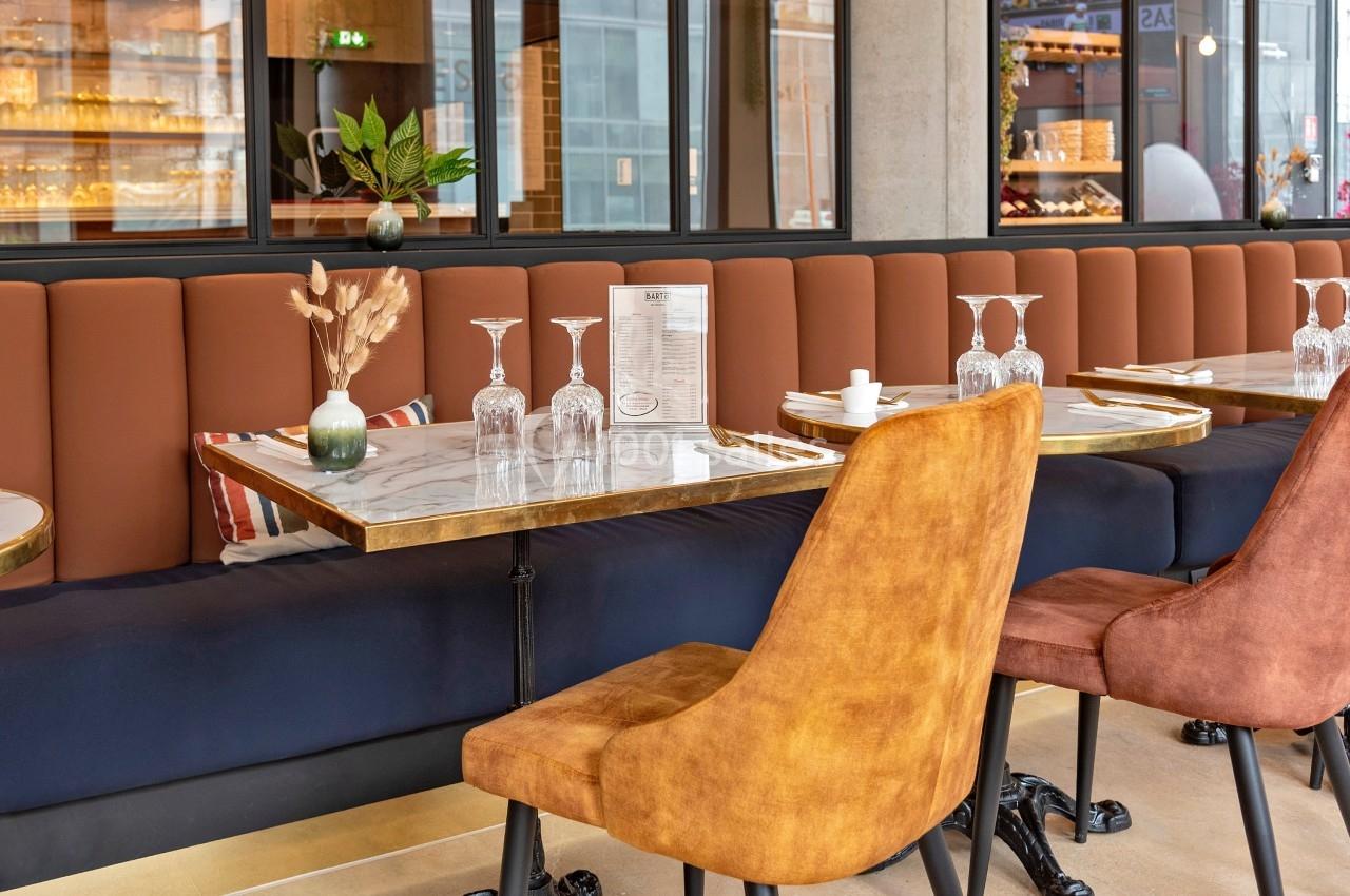 Salle de restaurant moderne avec tables en marbre, chaises rembourrées et banquette orange, décorée de plantes et de verres.