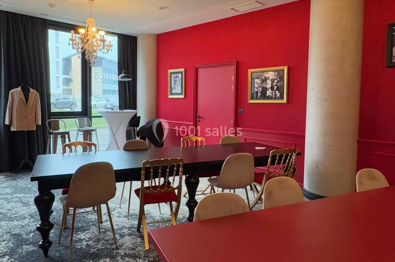 Salle de réunion avec murs rouges, table noire entourée de chaises, lustre et lumière naturelle provenant d'une baie vitrée.