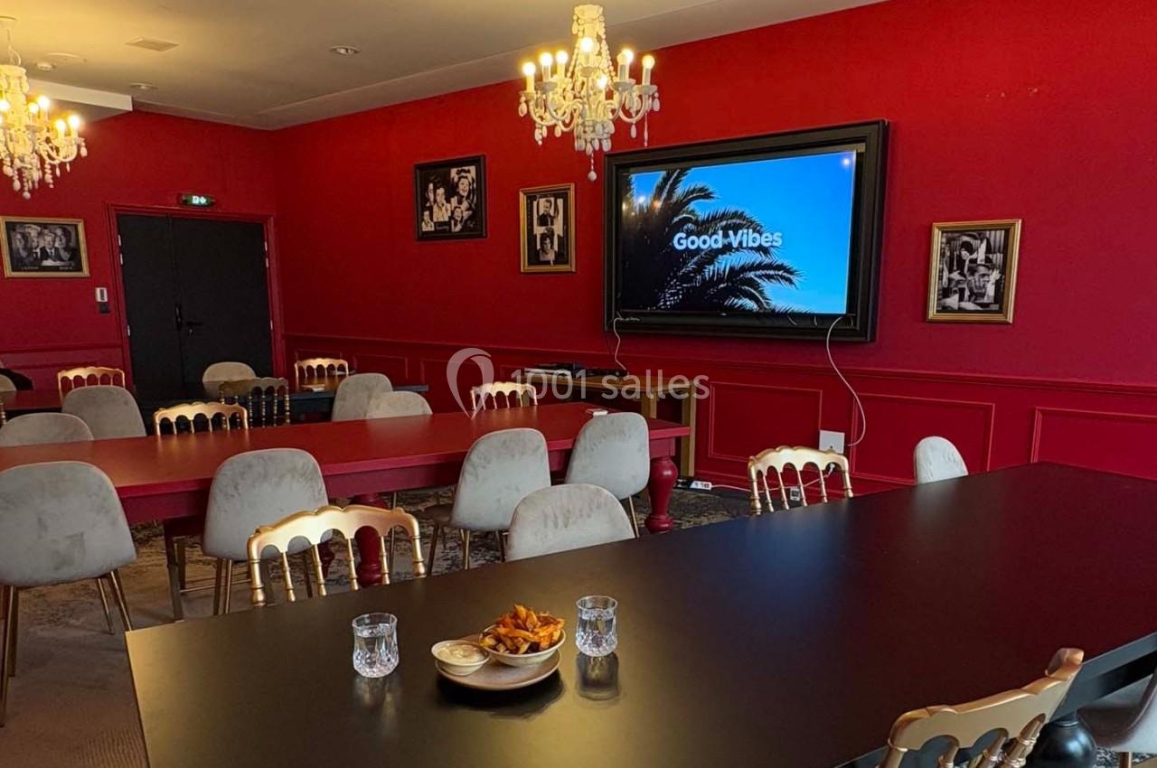 Salle de réunion avec murs rouges, tables noires et rouges, chaises beige, écran affichant ’Good Vibes’, et lustres…