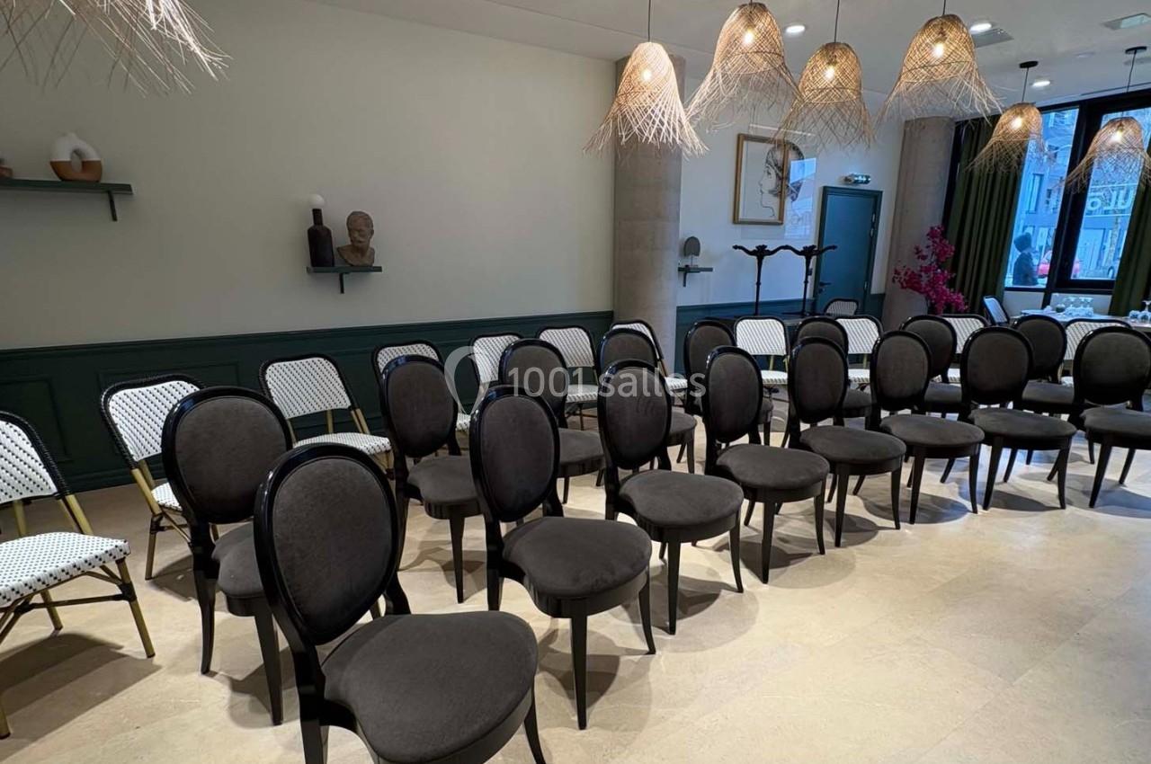 Salle aménagée avec des rangées de chaises noires et des luminaires suspendus dans un décor sobre et moderne.