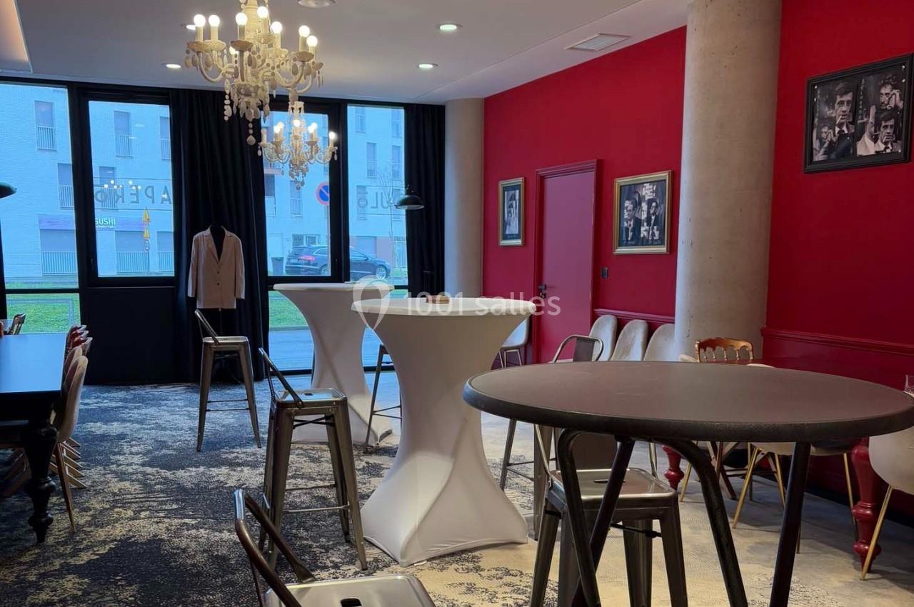 Salle avec murs rouges, tables hautes, chaises métalliques, lustre et photos encadrées, éclairée par des fenêtres.