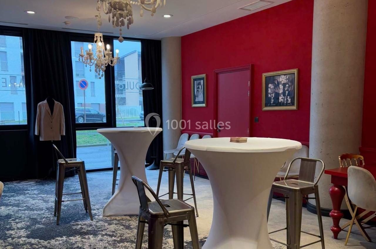Salle aménagée avec des tables hautes, des chaises en métal, des murs rouges décorés de cadres et un éclairage par lustres.