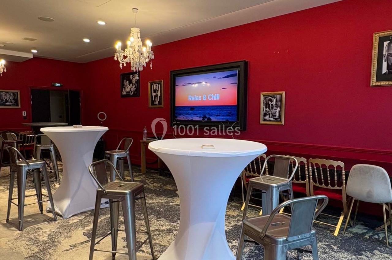 Salle avec murs rouges, tables hautes drapées de blanc, chaises variées et écran affichant ’Relax & Chill’.