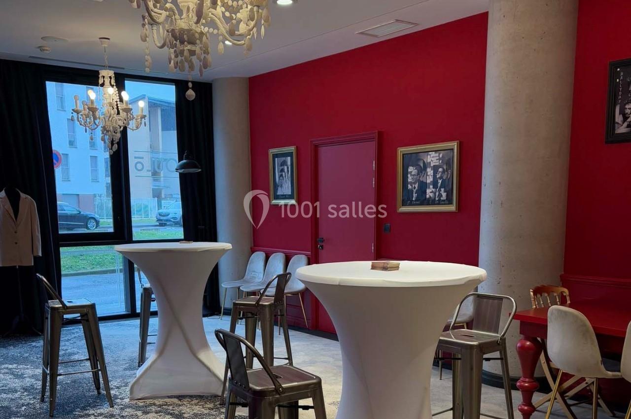 Salle moderne avec murs rouges, tables hautes blanches, chaises métalliques, lustres et cadres décoratifs.