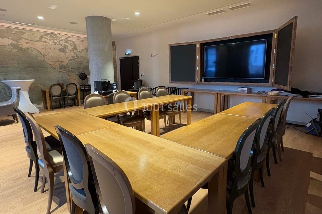 Salle de réunion avec des tables en bois disposées en U, des chaises, un écran mural et une carte ancienne en arrière-plan.