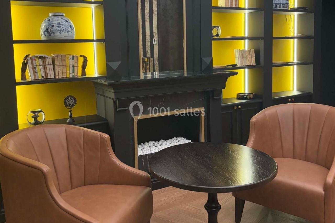 Deux fauteuils en cuir marron entourent une table ronde devant une cheminée encadrée de bibliothèques rétroéclairées.