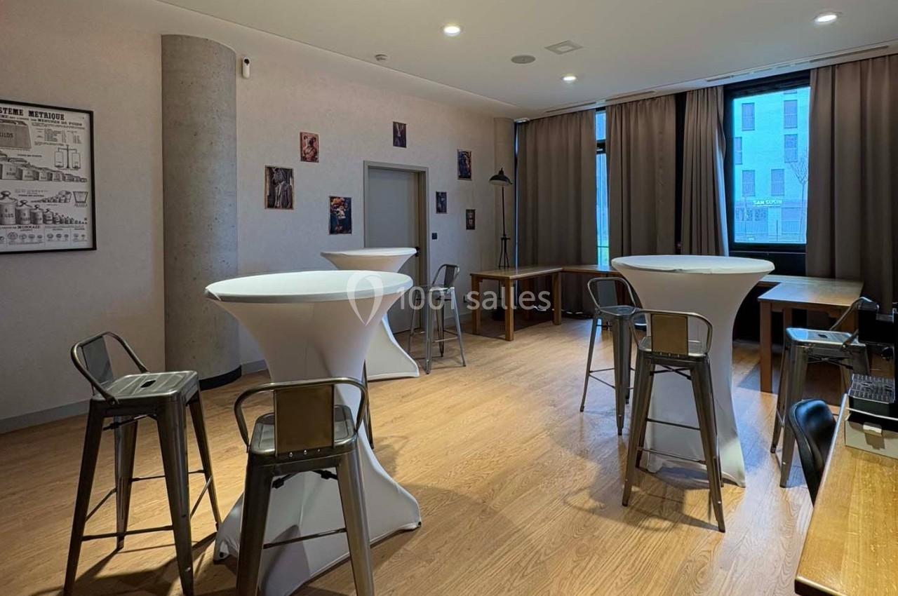 Salle lumineuse avec tables hautes, tabourets métalliques, parquet clair et grandes fenêtres avec rideaux.