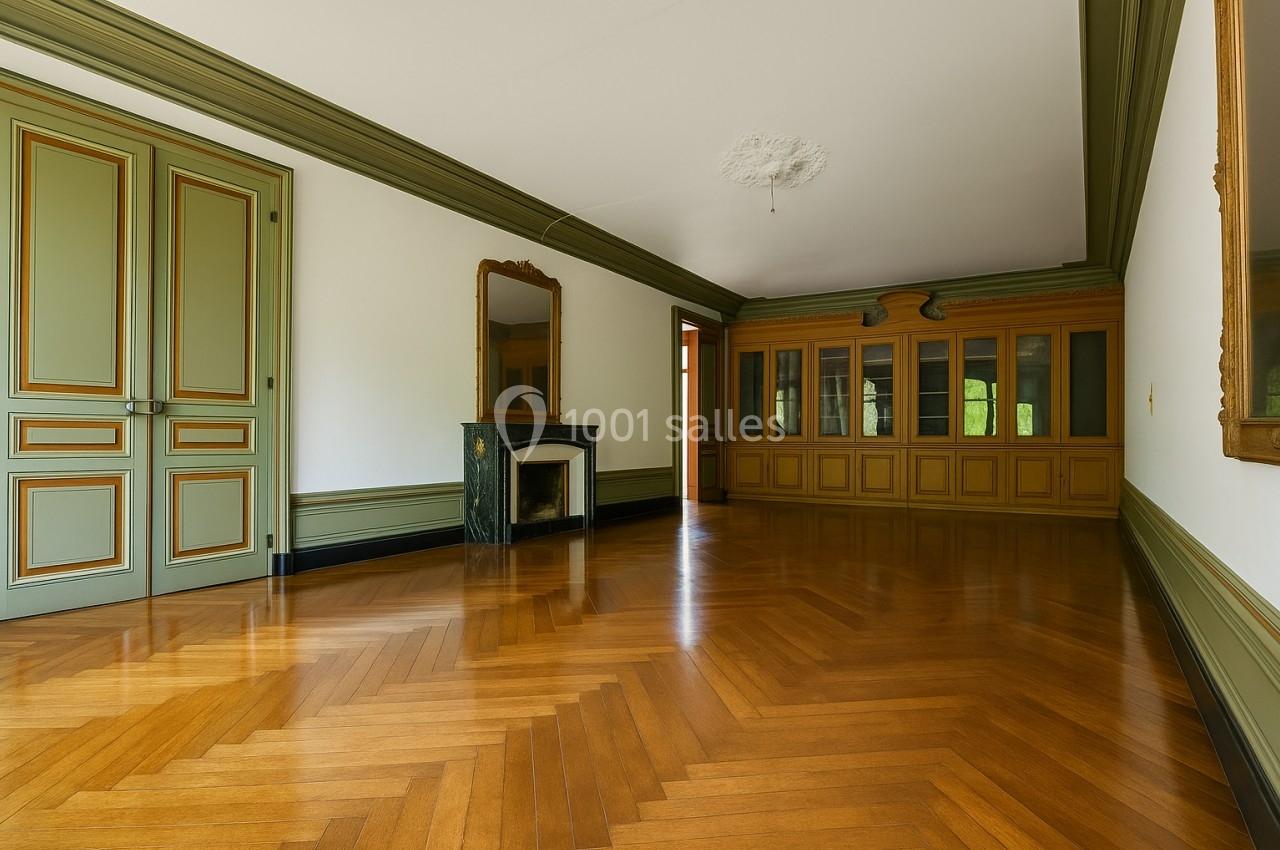 Salle vide avec parquet en chevrons, cheminée en marbre, miroir, boiseries vertes et grandes portes vitrées intégrées.