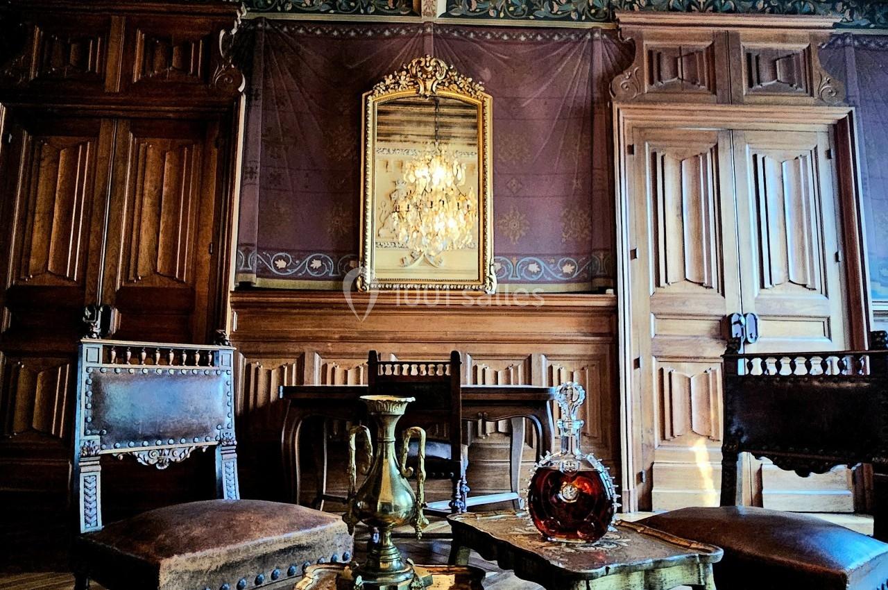 Salle avec mobilier ancien, tables en bois sculpté, chandelier en cristal et boiseries murales ornées.