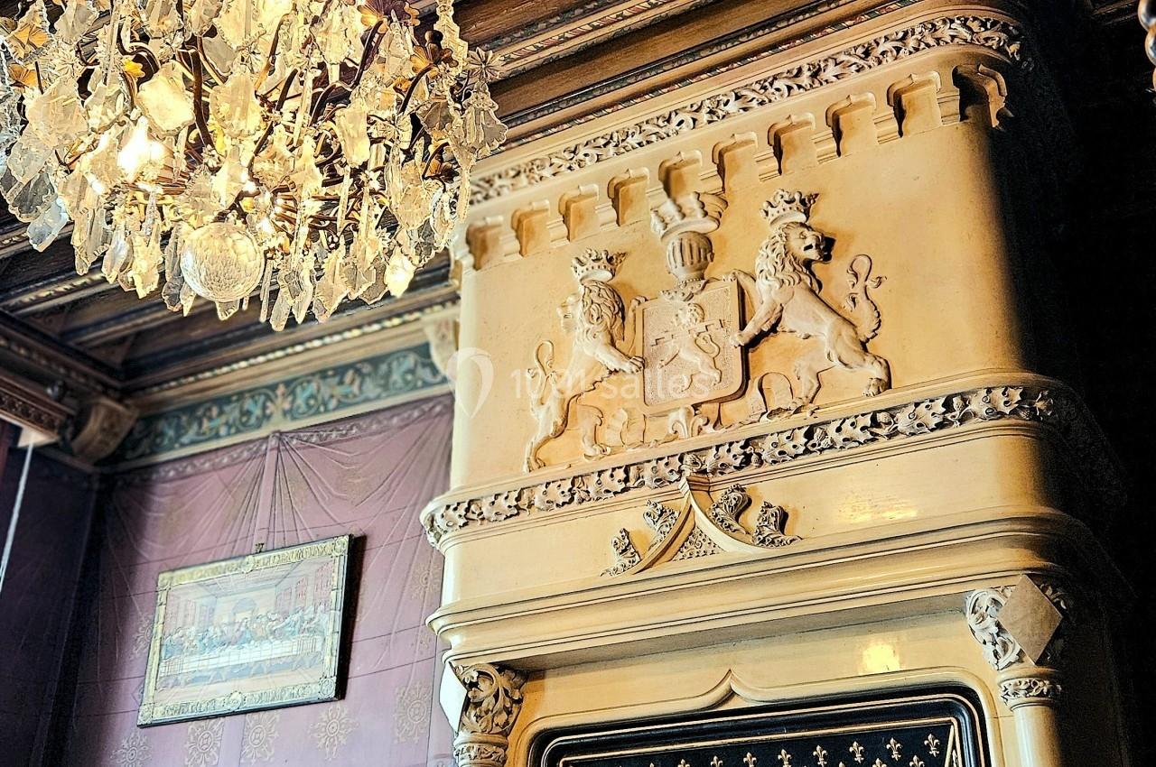 Cheminée sculptée ornée de motifs héraldiques, sous un plafond en bois décoré et un lustre en cristal.