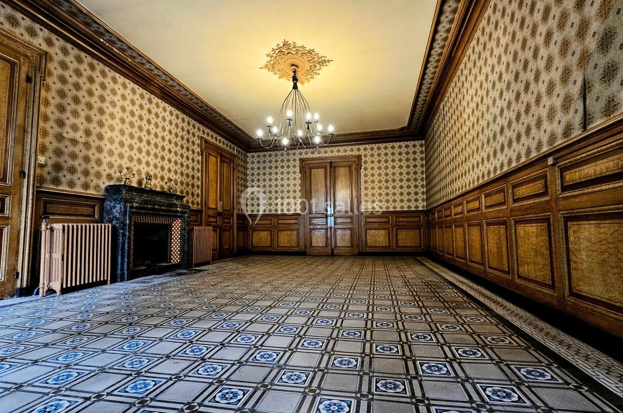Salle vide avec carrelage ancien à motifs, murs ornés de boiseries et papier peint, cheminée et lustre central.