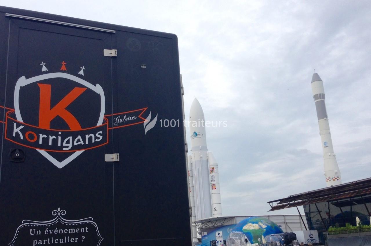 Camion marqué ’Korrigans’ devant des fusées Ariane exposées sous un ciel nuageux.
