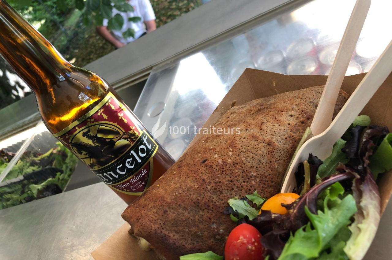 Bouteille de bière artisanale, galette de sarrasin et salade colorée dans un contenant en carton sur une table extérieure.