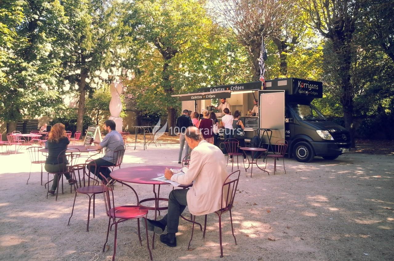 Un food truck servant des crêpes dans un parc avec des tables et des clients assis ou en file d'attente.