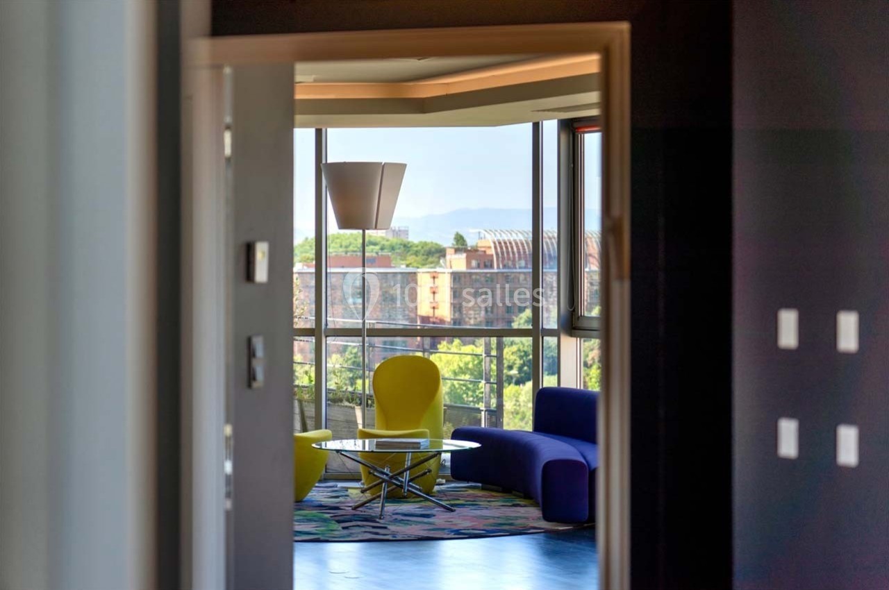 Location salle Caluire-et-Cuire (Rhône) - Le Belvédère #14 Vue d'un salon moderne avec fauteuils colorés, table basse et grande baie vitrée donnant sur un paysage urbain.