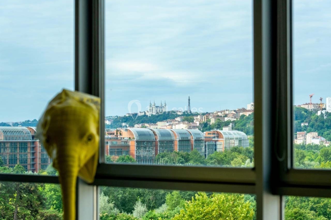 Location salle Caluire-et-Cuire (Rhône) - Le Belvédère #15 Vue sur des bâtiments modernes, des collines et la basilique de Fourvière à travers une fenêtre, avec une sculpture en…