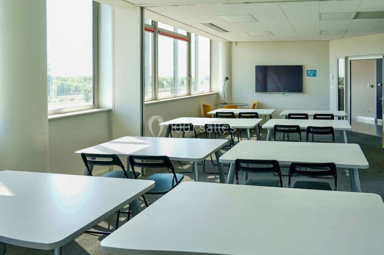 Location salle Caluire-et-Cuire (Rhône) - Le Belvédère #13 Salle de classe lumineuse avec des tables blanches, des chaises noires, un écran mural et de grandes fenêtres.