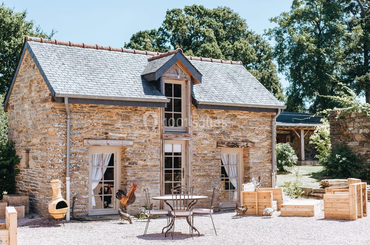 Maison en pierre avec toit en ardoise, jardin aménagé, table et chaises en métal, et coqs décoratifs devant.