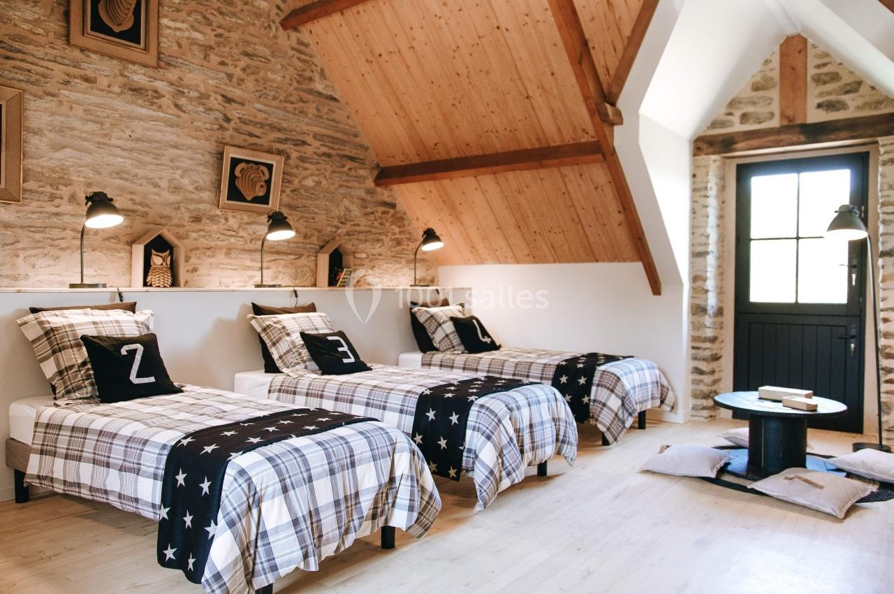 Chambre avec trois lits simples alignés, murs en pierre apparente, plafond en bois et lumière naturelle entrant par une…