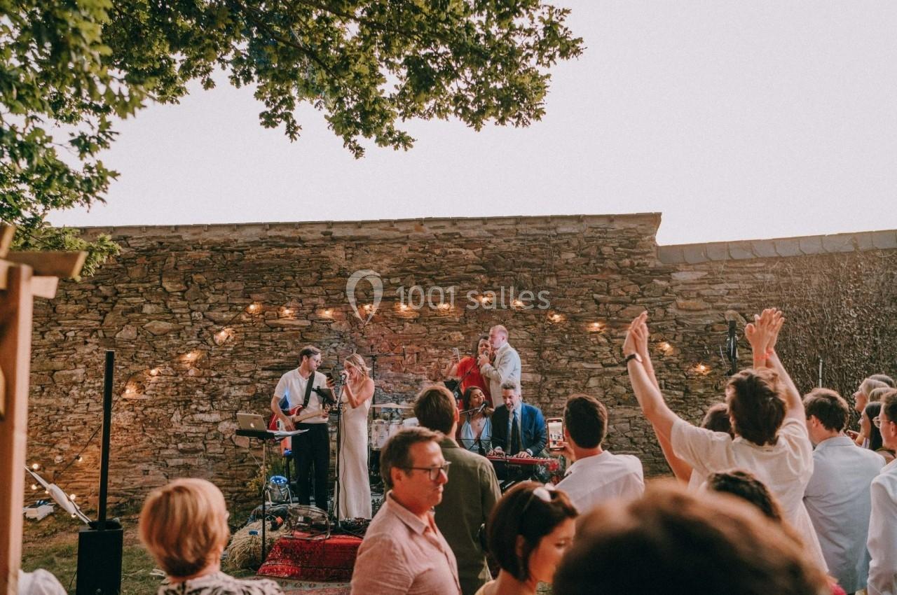Un groupe de musiciens joue en plein air devant un mur en pierre, entouré d'un public enthousiaste sous un ciel clair.