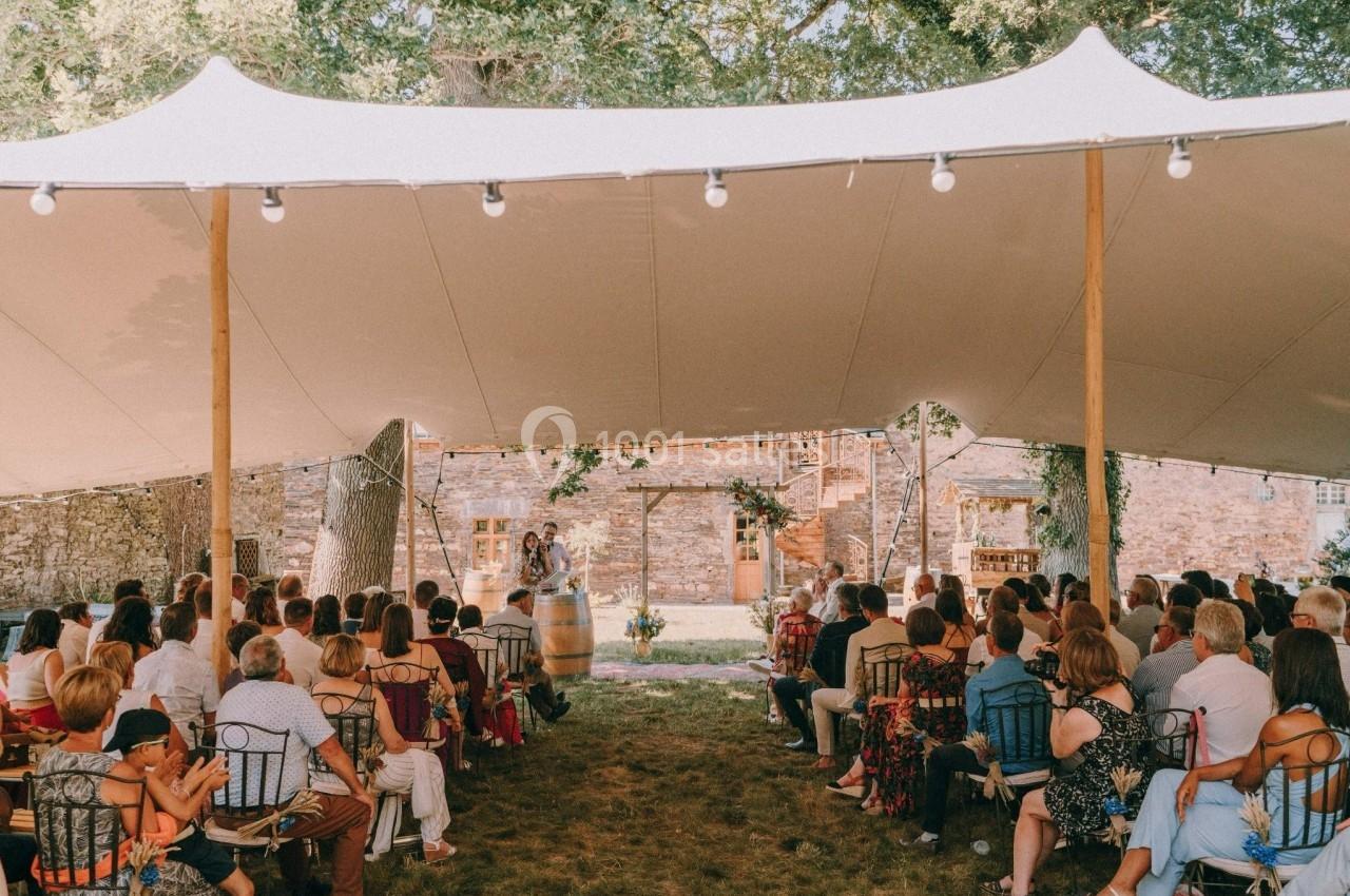 Cérémonie en plein air sous une tente, avec des invités assis et un couple debout devant un bâtiment en pierre.