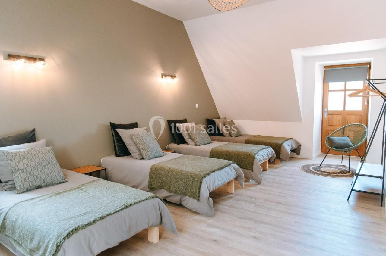 Quatre lits simples alignés dans une chambre lumineuse avec parquet, murs beiges et un fauteuil près d'une porte.