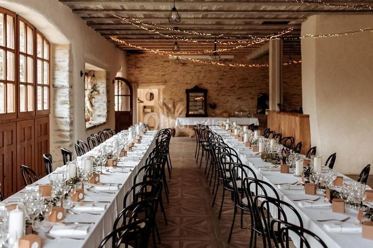 Salle de réception décorée avec de longues tables dressées, chaises noires et guirlandes lumineuses suspendues au plafond.