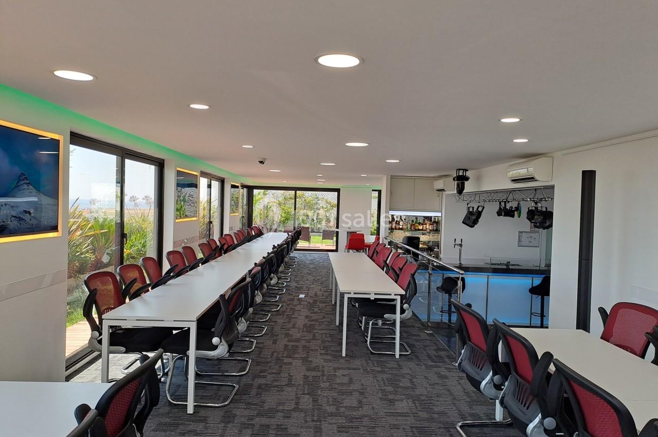 Salle de réunion lumineuse avec de longues tables, des chaises rouges et un bar en arrière-plan.
