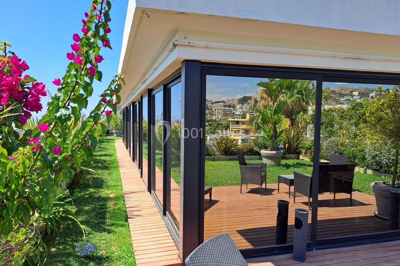 Terrasse en bois avec mobilier extérieur, entourée de verdure et de fleurs, jouxtant une maison aux grandes baies vitrées.