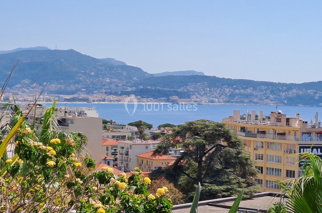Vue sur une ville côtière avec des bâtiments, des arbres et des montagnes en arrière-plan, bordée par la mer.