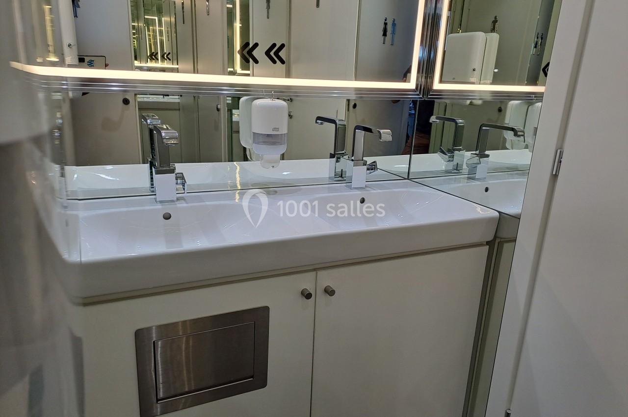 Double lavabo moderne avec robinets chromés, distributeur de savon mural et grand miroir éclairé en arrière-plan.