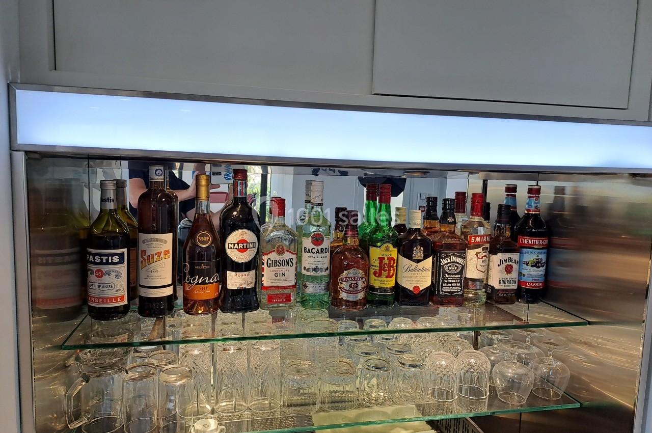 Bouteilles d'alcools variés alignées sur des étagères en verre, avec des verres disposés en dessous.