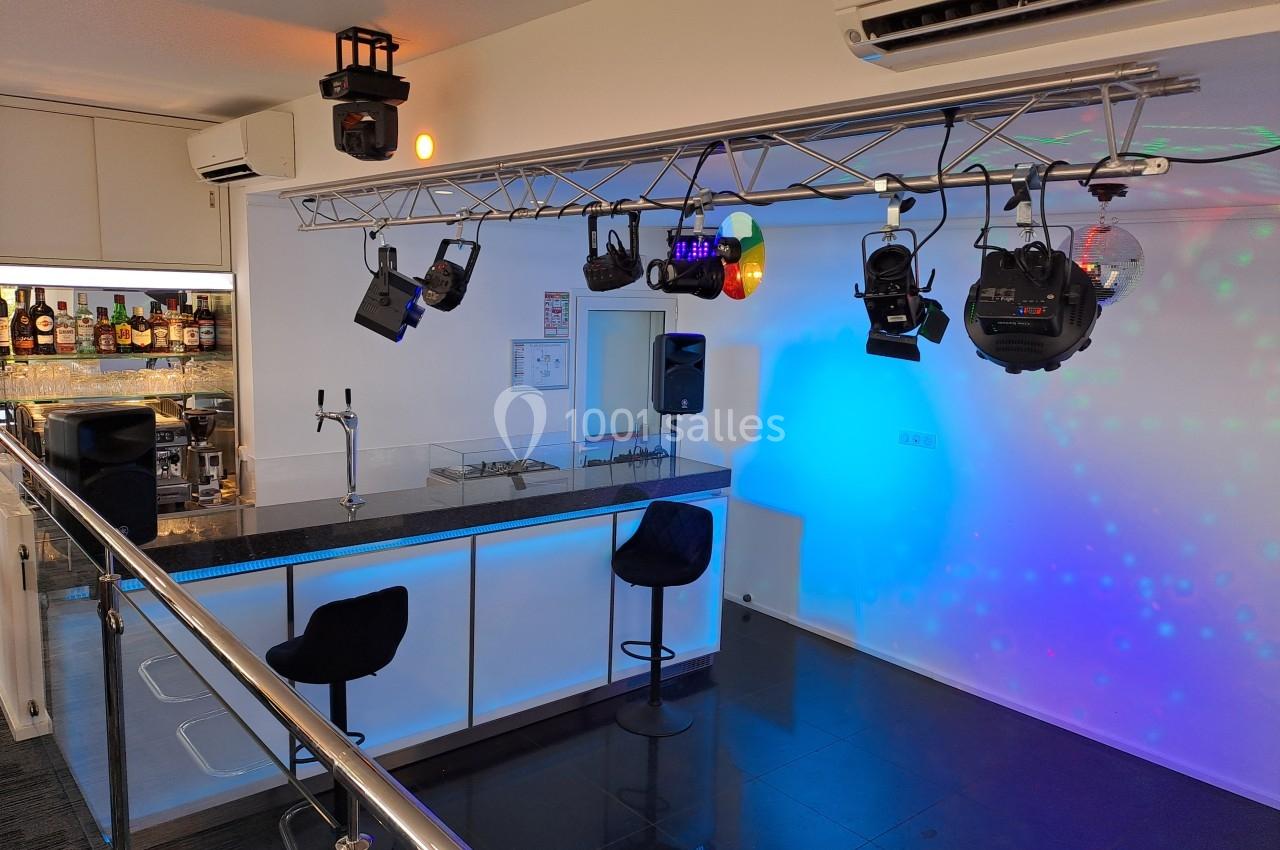 Bar moderne avec comptoir éclairé, tabourets noirs, projecteurs suspendus et éclairage coloré dans une salle.