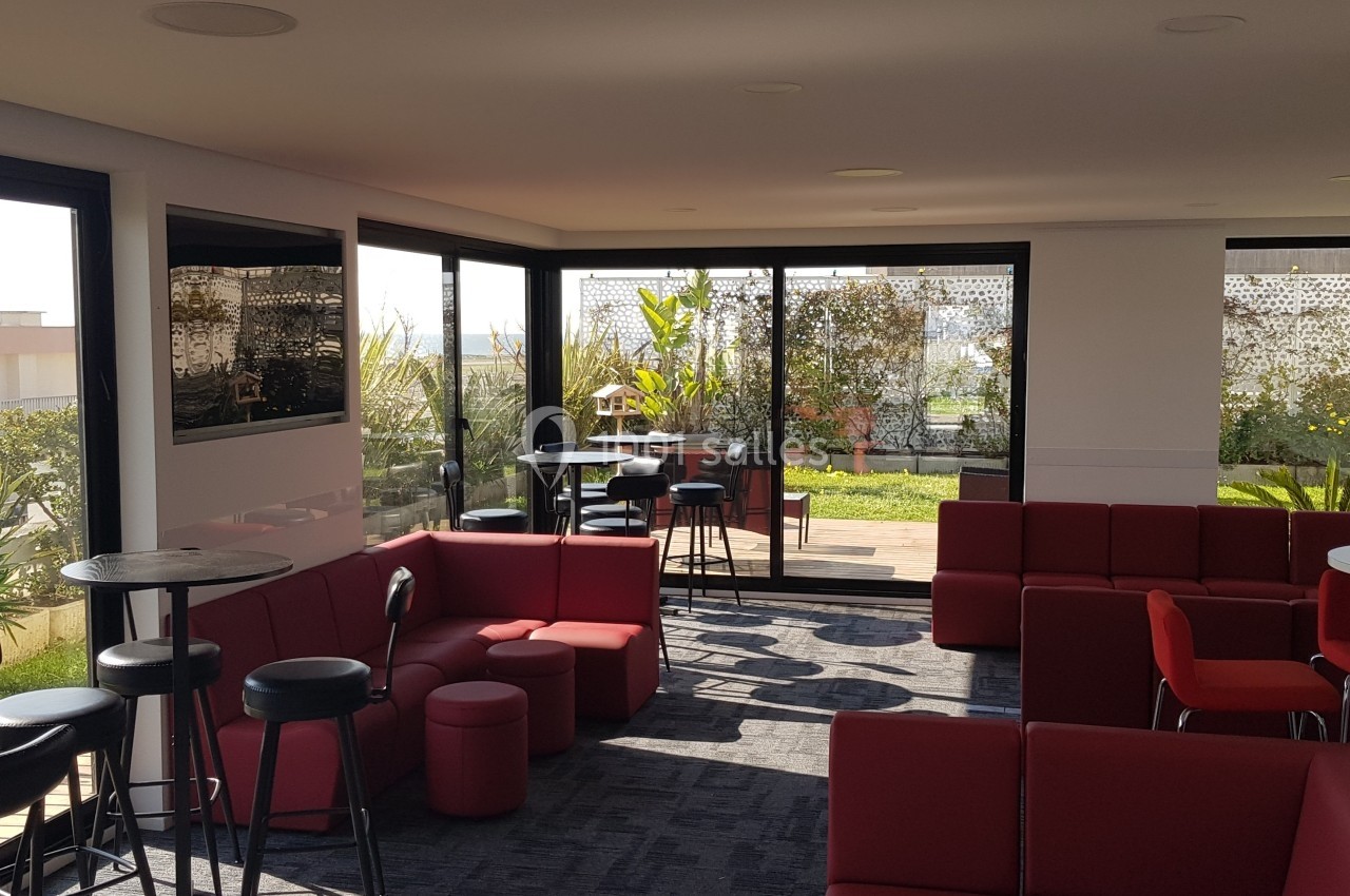 Espace intérieur lumineux avec canapés rouges, tables hautes et vue sur une terrasse végétalisée.