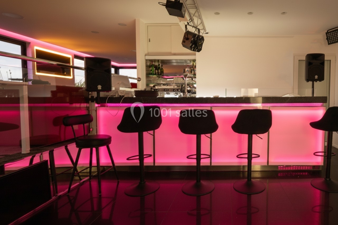 Bar moderne avec comptoir éclairé en rose, tabourets noirs alignés et équipement audio visible en arrière-plan.