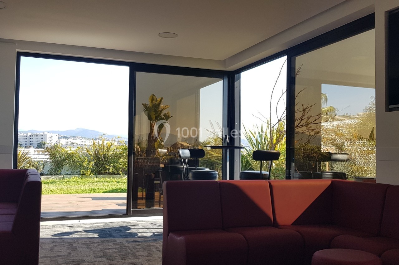 Espace lounge lumineux avec canapés rouges, grandes baies vitrées donnant sur une terrasse et vue sur des montagnes.