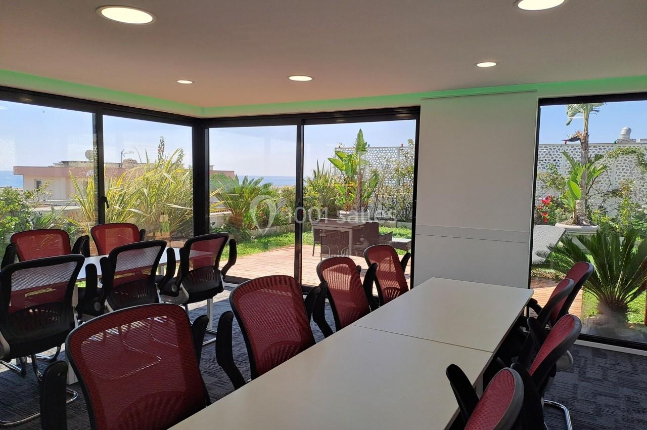 Salle de réunion lumineuse avec des chaises rouges, une grande table et vue sur un jardin extérieur.