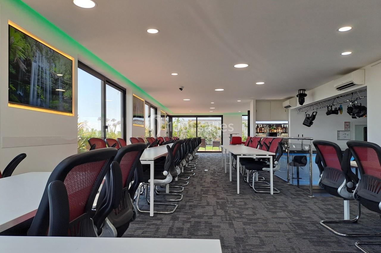 Salle de réunion moderne avec des chaises noires et rouges, tables blanches, éclairage LED et grandes fenêtres.